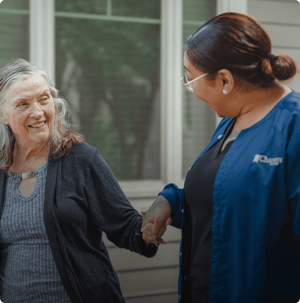 An older woman smiles while holding hands with a caregiver in a blue uniform, standing outdoors in front of a building with large windows. An older woman smiles while holding hands with a caregiver in a blue uniform, standing outdoors in front of a building with large windows.