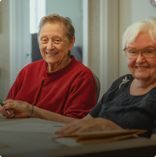 Two elderly women sit at a table, smiling and holding pens, appearing to enjoy a shared activity in a well-lit room. Two elderly women sit at a table, smiling and holding pens, appearing to enjoy a shared activity in a well-lit room.